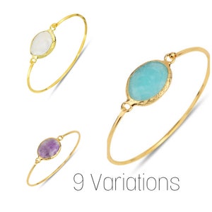 May include: Three gold bangle bracelets with different colored oval-shaped gemstones. The bracelets are shown on a white background. The gemstones are white, light blue, and purple.