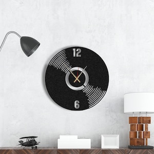 Peut inclure: Une horloge en forme de disque vinyle noir et blanc avec des chiffres et des aiguilles argentés. L'horloge est accrochée à un mur blanc au-dessus d'un meuble en bois sur lequel se trouve une petite figurine d'avion.