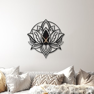 Puede incluir: Un reloj de pared blanco y negro con un diseño de flor de loto. El reloj tiene manecillas doradas y está colgado en una pared blanca sobre un sofá blanco con almohadas.