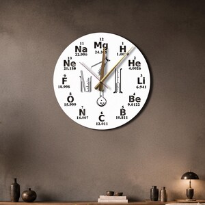 May include: A round wall clock with a white face featuring black chemical element symbols and atomic numbers from the periodic table arranged in a circle. The center displays black line art of laboratory glassware and gold clock hands.