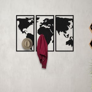 May include: A black metal wall-mounted coat rack shaped like a world map. The rack has three sections and is mounted on a white wall. A red jacket and a brown hat are hanging on the rack.