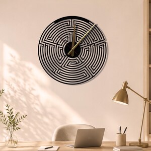 Puede incluir: Un reloj de pared circular con un patrón de laberinto en blanco y negro en su esfera. El reloj tiene dos manecillas delgadas de metal dorado posicionadas en el centro. El diseño consiste en líneas laberínticas concéntricas y complejas.