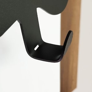 May include: Black metal hook attached to a wooden wall mount. The hook is shaped like a stylized animal head with a rounded, protruding hook at the bottom.