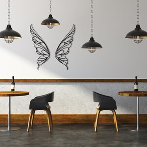K&ouml;nnte beinhalten: Wandkunst aus schwarzen Metall-Schmetterlingsfl&uuml;geln. Das dekorative St&uuml;ck ist an einer wei&szlig;en Wand angebracht. Die Fl&uuml;gel sind symmetrisch und weisen ein kompliziertes, offenes Design auf. Der Raum hat schwarze Pendelleuchten und Bistrotische.