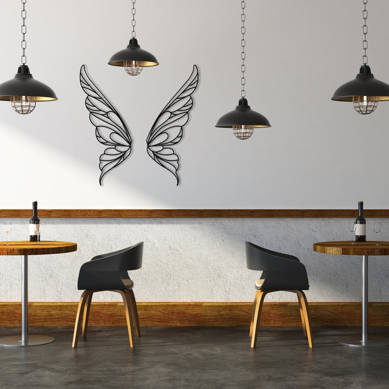 K&ouml;nnte beinhalten: Wandkunst aus schwarzen Metall-Schmetterlingsfl&uuml;geln. Das dekorative St&uuml;ck ist an einer wei&szlig;en Wand angebracht. Die Fl&uuml;gel sind symmetrisch und weisen ein kompliziertes, offenes Design auf. Der Raum hat schwarze Pendelleuchten und Bistrotische.