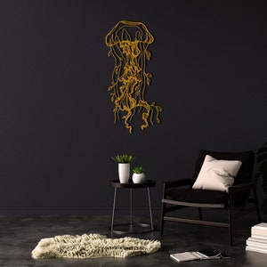 May include: A gold metal wall sculpture of a jellyfish with intricate details. The sculpture is hanging on a black wall in a room with a black armchair and a side table with two potted plants.