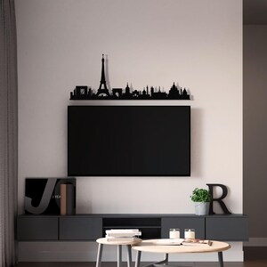 May include: Black metal Paris skyline wall decor with the Eiffel Tower as the centerpiece. The decor is mounted above a black television on a gray wall. The television is on a gray entertainment center with a black book and a green plant on the shelf below.