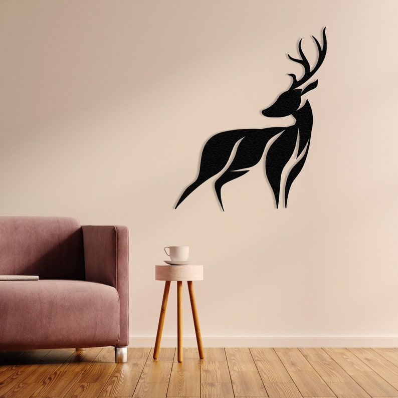 May include: Black silhouette of a deer with antlers, mounted on a light beige wall. The deer is facing right and has a stylized, abstract design. A pink side table with a white cup sits in front of a pink sofa.
