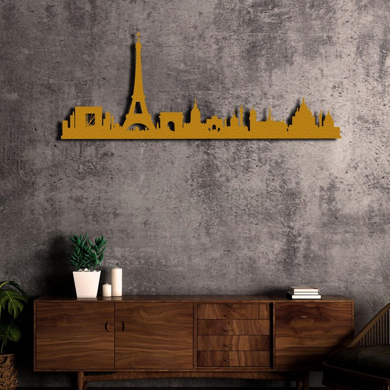 May include: A gold metal wall decor featuring a silhouette of the Paris skyline, including the Eiffel Tower, Arc de Triomphe, and other iconic landmarks. The decor is mounted on a gray concrete wall above a wooden dresser.