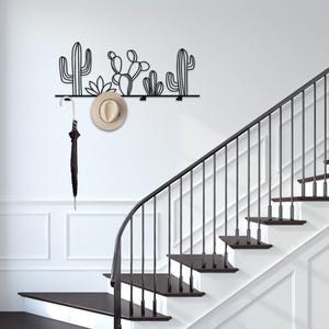 May include: Black metal wall mounted coat rack with a cactus design. A hat and umbrella are hanging on the rack. The rack is mounted on a white wall with a black staircase in the background.