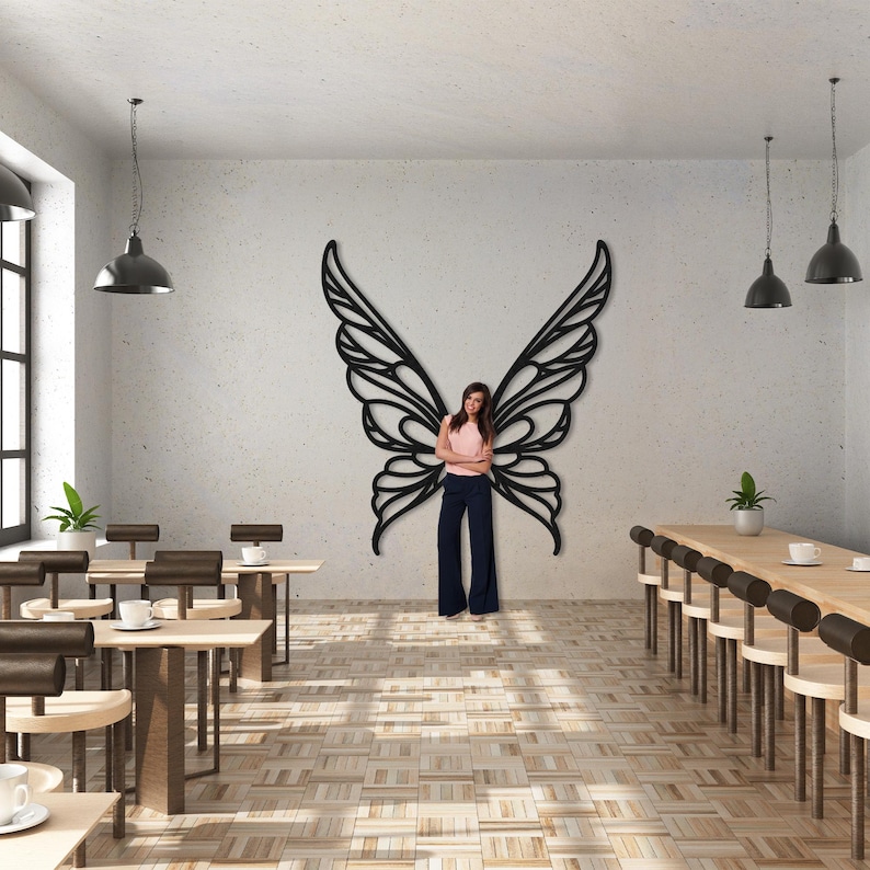 May include: A large, black metal butterfly wing wall art decor. The wings are intricately designed with a delicate, openwork pattern. The artwork is mounted on a white wall in a room with tables and chairs, and pendant lights.