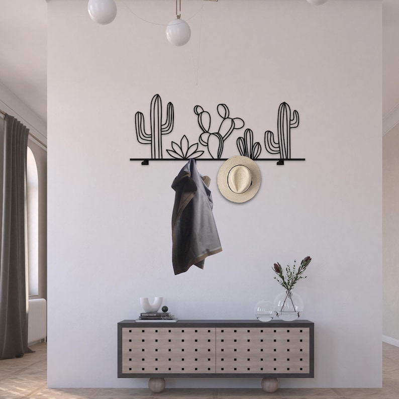 May include: Black metal wall-mounted coat rack with a cactus design. The rack has five hooks and is mounted on a white wall. A hat and a jacket are hanging on the hooks.
