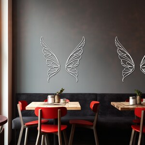 K&ouml;nnte beinhalten: Wei&szlig;e, lasergeschnittene Wandkunst in Form von Schmetterlingsfl&uuml;geln. Die Fl&uuml;gel werden paarweise mit filigranen Details und einem zarten, luftigen Design dargestellt. Sie sind an einer dunkelgrauen Wand angebracht und bilden einen auff&auml;lligen Kontrast.