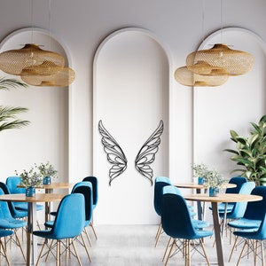 K&ouml;nnte beinhalten: Schwarze Metall-Wandkunst mit Schmetterlingsfl&uuml;geln. Die Fl&uuml;gel sind an einer wei&szlig;en Wand in einem Restaurant angebracht. Das Restaurant hat blaue St&uuml;hle, Holztische und gewebte Pendelleuchten.