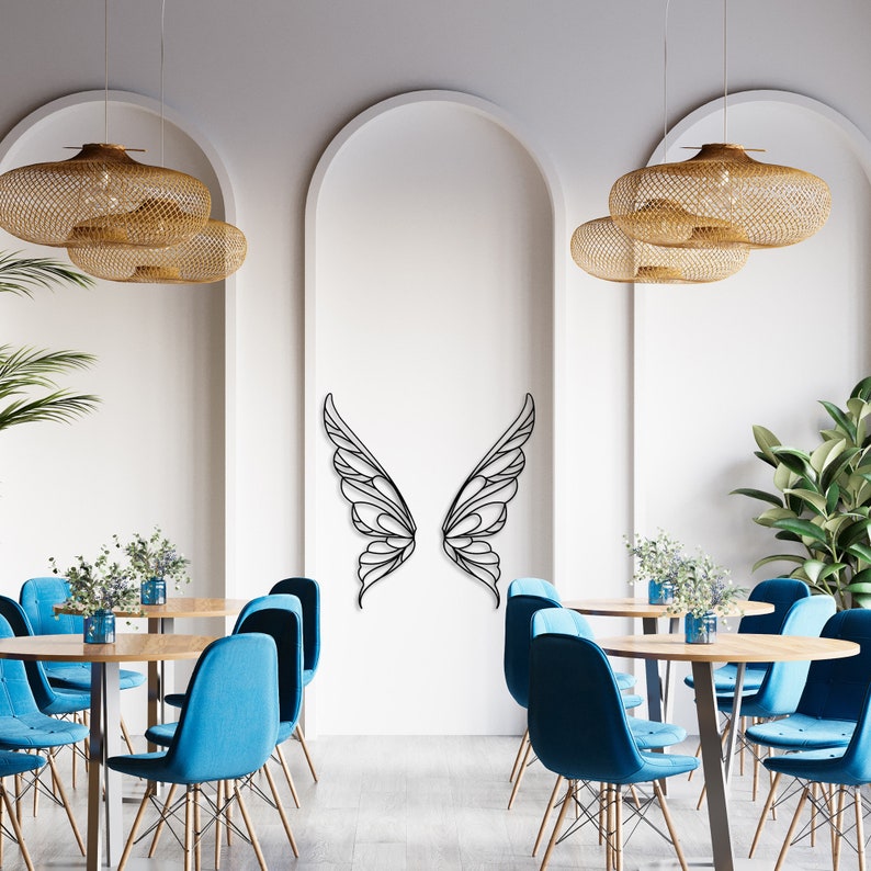 K&ouml;nnte beinhalten: Schwarze Metall-Wandkunst mit Schmetterlingsfl&uuml;geln. Die Fl&uuml;gel sind an einer wei&szlig;en Wand in einem Restaurant angebracht. Das Restaurant hat blaue St&uuml;hle, Holztische und gewebte Pendelleuchten.