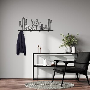 May include: Black metal wall mounted coat rack with a cactus design. The rack has five hooks and is mounted on a white wall. A black chair and a black shelf with books are in the background.