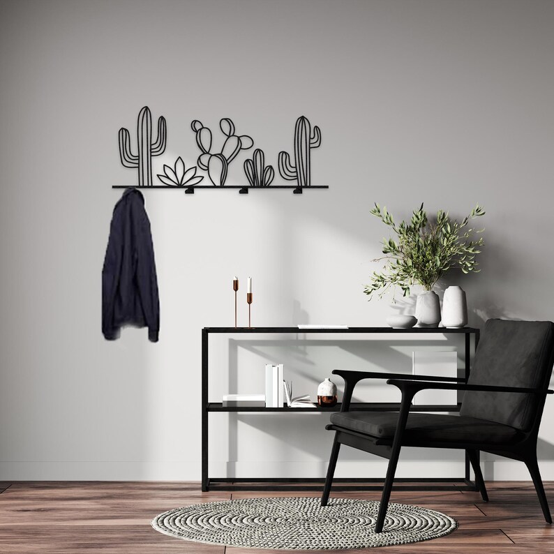 May include: Black metal wall mounted coat rack with a cactus design. The rack has five hooks and is mounted on a white wall. A black chair and a black shelf with books are in the background.