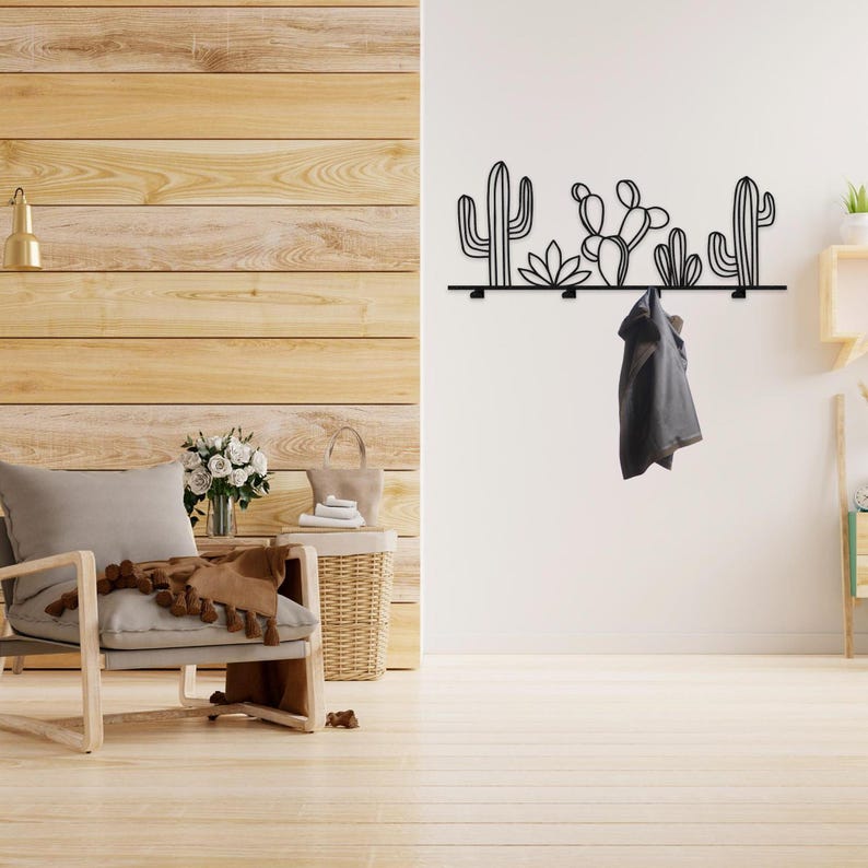 May include: Black metal wall-mounted coat rack with a cactus design. The rack has five hooks and is mounted on a white wall. A gray jacket is hanging on one of the hooks.
