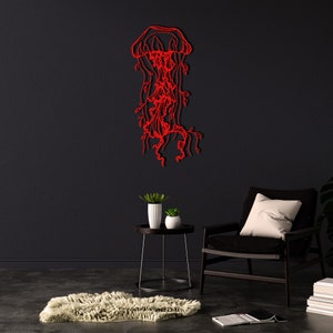 May include: Red metal wall art sculpture of a jellyfish with tentacles and flowing tendrils. The sculpture is mounted on a black wall in a room with a black armchair and a small side table with plants.