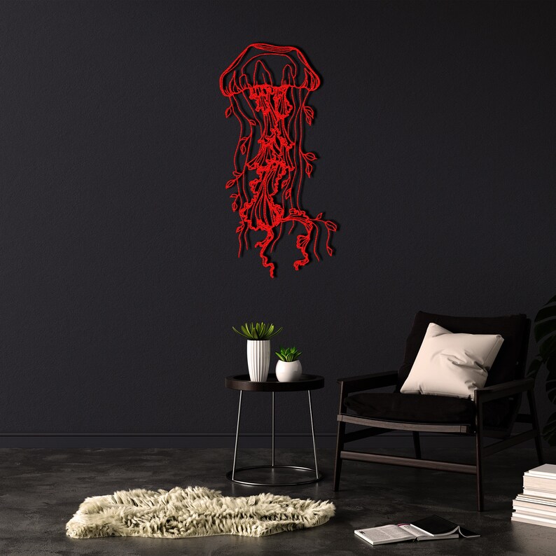 May include: Red metal wall art sculpture of a jellyfish with tentacles and flowing tendrils. The sculpture is mounted on a black wall in a room with a black armchair and a small side table with plants.