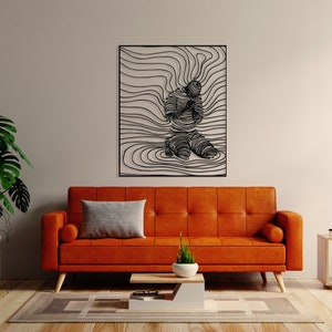 Op de afbeelding: Een zwarte metalen wandsculptuur die een persoon in een knielende houding voorstelt, omlijnd met golvende lijnen. De sculptuur hangt aan een muur boven een oranje bank.