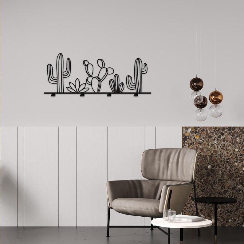 May include: Black metal wall decor featuring a row of cacti and succulents. The decor is mounted on a white wall with a brown leather armchair and a small table in the foreground.