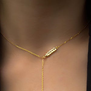 Puede incluir: Un delicado collar de oro con un colgante en forma de espiga de trigo. El colgante está centrado en una fina cadena, con una cadena más larga que se extiende hacia abajo. El collar se lleva sobre un fondo oscuro, resaltando el brillo del oro.