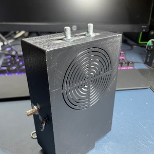 May include: A black box with a circular speaker grill and two silver knobs on top. The box has a small silver knob on the side and a spring attached to a metal ring.