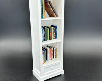 May include: White wooden bookcase with three shelves and a drawer. The bookcase is filled with a variety of books, including some with colorful covers.