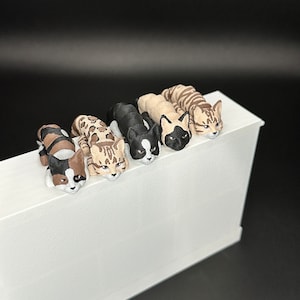 May include: Five small, 3D printed cat figurines in various colors and patterns, including black and white, brown tabby, and calico. The figurines are arranged in a row on a white surface, with their heads facing the viewer.