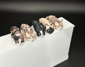 May include: Five small, 3D printed cat figurines in various colors and patterns, including black and white, brown tabby, and calico. The figurines are arranged in a row on a white surface, with their heads facing the viewer.