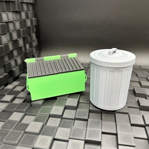 May include: A green and black miniature dumpster and a gray miniature trash can. Both are made of plastic and are sitting on a black tiled surface.