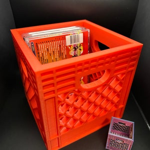 May include: A red plastic crate with a CD case inside. The crate has a handle on the side and a grid pattern. There is a smaller red plastic crate in the background.