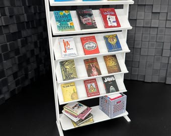 May include: White bookshelf with five shelves, each filled with miniature books. The books have various titles, including "The Shape of Things to Come", "Diary of a Wimpy Kid", "Jaws", and "The Little Prince". A gray crate sits on the bottom shelf.