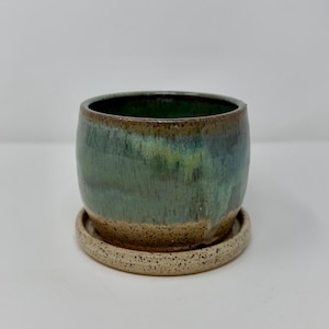 May include: A cylindrical ceramic planter with a smooth, glossy glaze in shades of deep forest green and teal that transitions into a speckled, textured beige base. The planter sits securely on a matching circular saucer with a speckled finish.