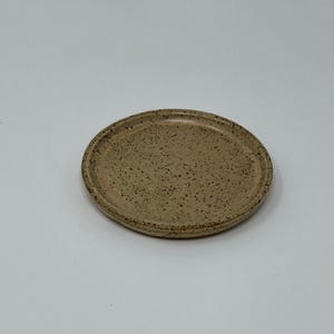 Puede incluir: Un plato pequeño, redondo, de cerámica beige moteado con un borde ligeramente elevado.