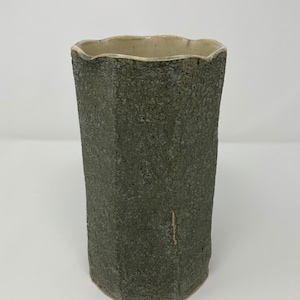 May include: A tall, textured ceramic vase with a dark gray exterior and a light beige interior. The vase has a unique, slightly irregular shape with a wavy rim. The surface has a rough, speckled appearance.