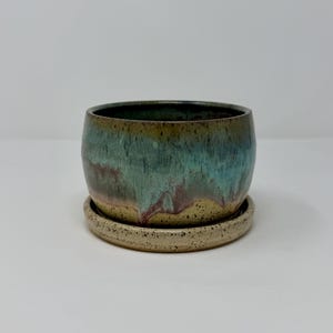 May include: A round ceramic planter with a speckled beige base and a reactive glaze featuring blended tones of teal, blue, and brown. The planter sits on a matching circular speckled beige saucer, creating a cohesive set for small plants.