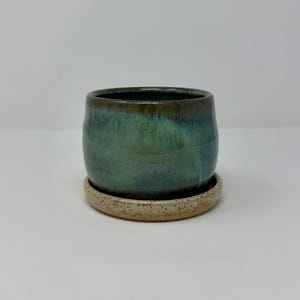 May include: A cylindrical ceramic planter with a glossy teal and dark green glaze that transitions into a deep brown rim. The planter sits on a matching circular saucer featuring a speckled, light beige, matte, and textured clay finish.