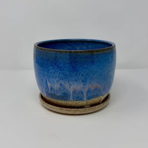 May include: A round ceramic planter with a glossy blue glaze that transitions into a speckled beige base. The planter features a matching circular saucer underneath, both displaying a textured, earthy finish with visible dark speckles.