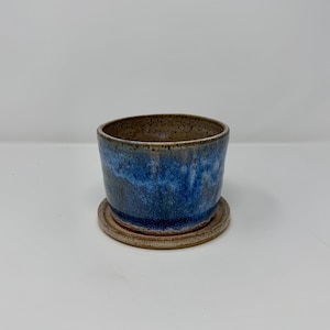 May include: A ceramic planter with a blue and brown glaze. The small, round pot sits on a matching saucer. The interior of the pot is brown, while the exterior features a blend of blue and brown hues.