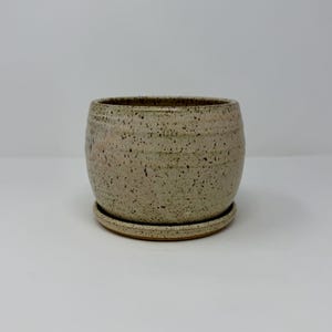 May include: A round ceramic planter with a speckled beige and light brown finish. The pot features a subtle horizontal ribbed texture along the exterior surface and sits securely on a matching circular ceramic saucer base.