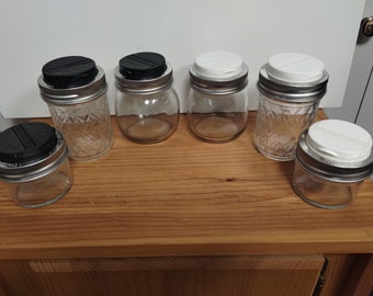 May include: Six glass jars with metal lids. Three jars have black lids and three jars have white lids. The jars are arranged in a row on a wooden surface.
