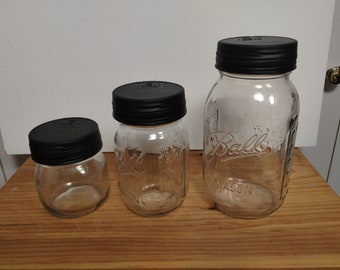 May include: Three glass jars with black lids. The jars are clear and have a textured surface. The largest jar has the word "Ball" and "Mason" printed on the side.
