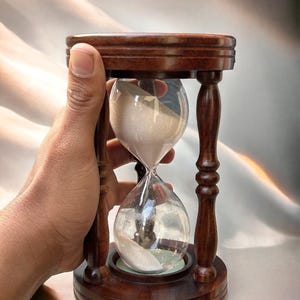 May include: A wooden hourglass with a dark brown finish. The hourglass is filled with white sand and is being held in a hand.