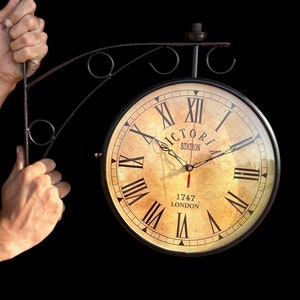 May include: A black metal clock with Roman numerals and a cream-colored face. The clock reads "Victoria Station 1747 London".