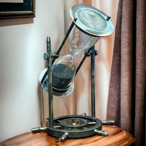 Puede incluir: Un reloj de arena vintage con arena negra, colocado sobre una mesa de madera con una base de rueda de barco. El reloj de arena está hecho de latón y vidrio y tiene una parte superior decorativa.