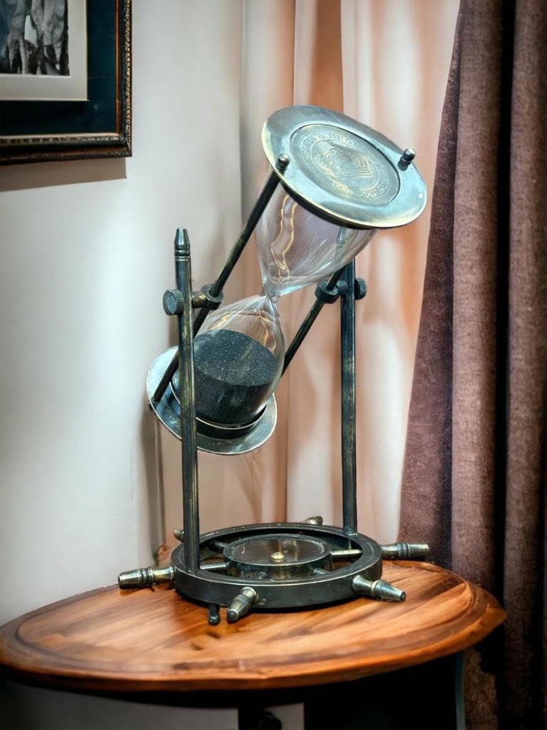 Op de afbeelding: Een vintage zandloper met zwart zand, geplaatst op een houten tafel met een scheepswielbasis. De zandloper is gemaakt van messing en glas en heeft een decoratieve bovenkant.