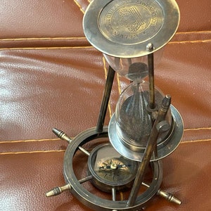 Op de afbeelding: Een zandloper van messing met een kompasvoet. De zandloper heeft een glazen bol en een metalen bovenkant met de inscriptie "RELI-VEN HUGHES LONDON 1817".