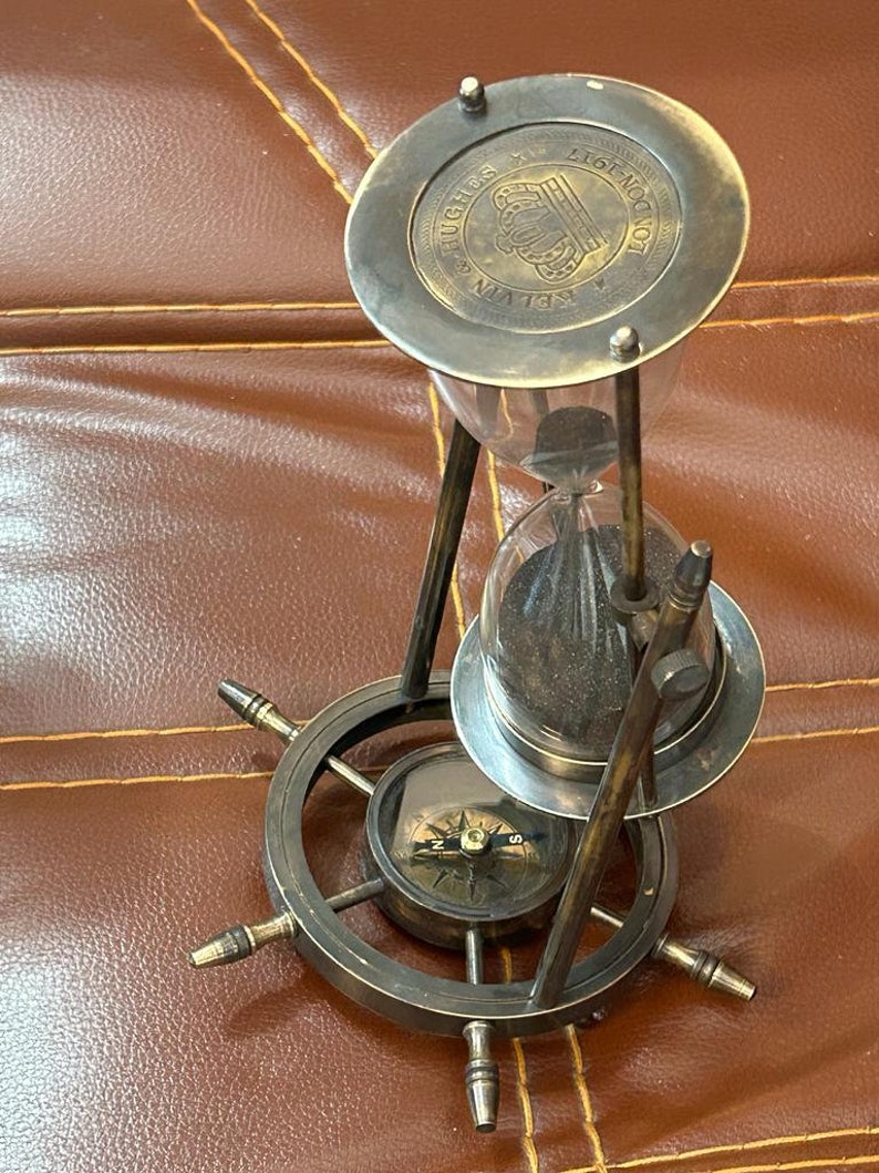 Op de afbeelding: Een zandloper van messing met een kompasvoet. De zandloper heeft een glazen bol en een metalen bovenkant met de inscriptie "RELI-VEN HUGHES LONDON 1817".